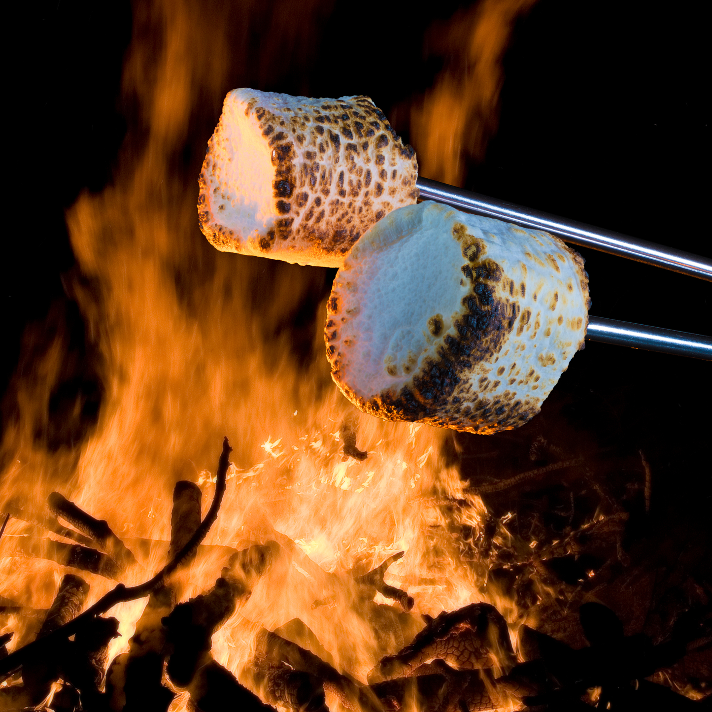 Two toasted marshmallows on metal skewers roasting over an open campfire at night, with bright orange flames and dark embers in the background.