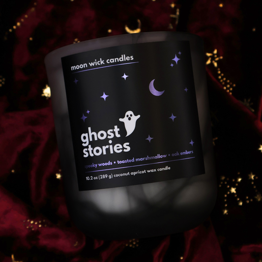 A black and white swirled glass jar candle from Moon Wick Candles, featuring a black label with metallic purple stars and a white ghost logo that reads 'ghost stories'. The candle is set against a burgundy velvet fabric background with gold celestial details.