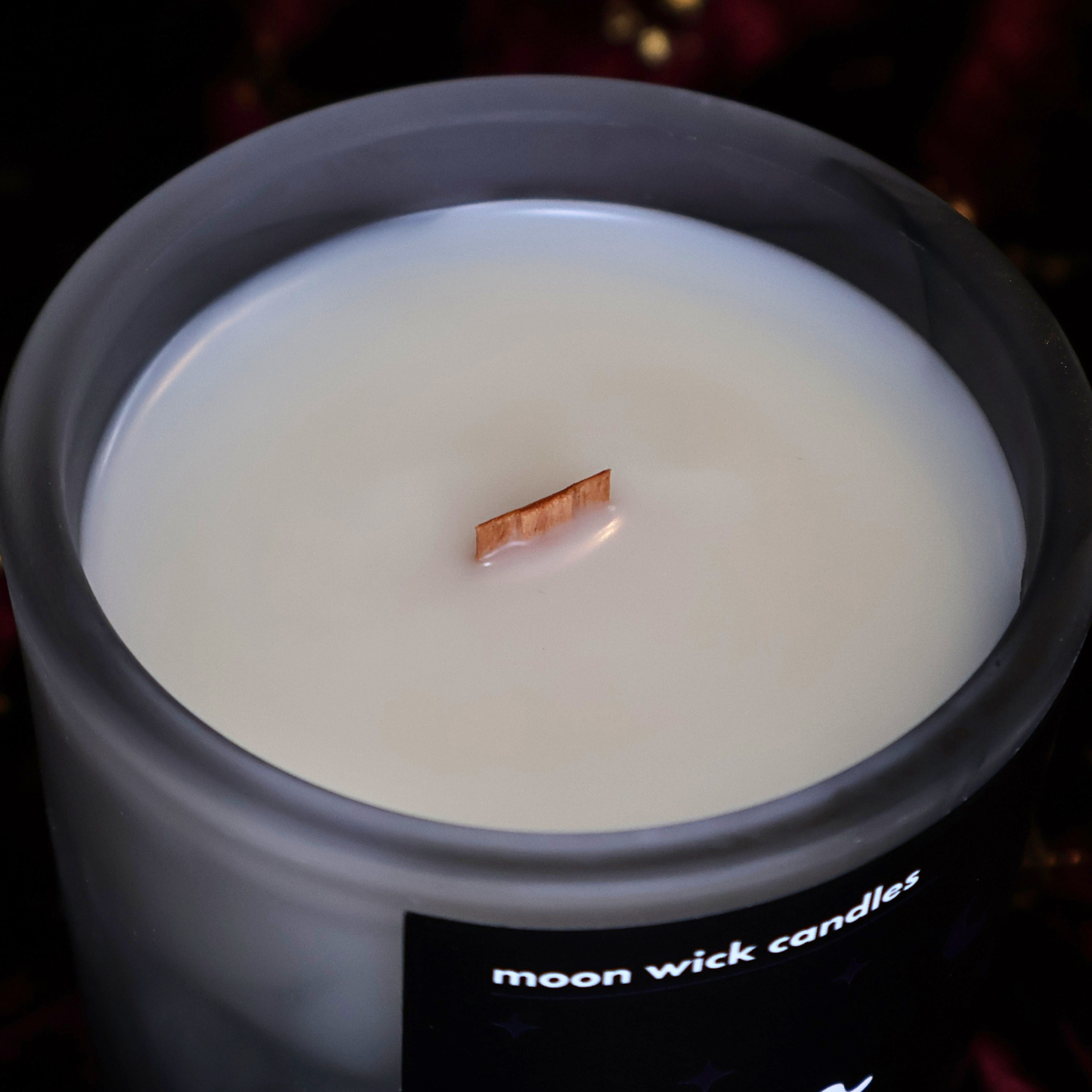 A close-up of a wax candle in a frosted glass jar with a wooden wick and a black label reading 'moon wick candles' on a dark background.