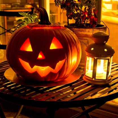 A glowing jack-o'-lantern pumpkin with carved facial features sits on a dark slatted table next to a lit lantern, with potted plants in the background.