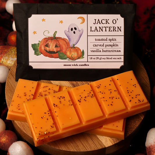 orange snap bar wax melt with orange glitter on top, arranged on a wooden surface in front of a black wax paper bag labeled 'Jack O' Lantern' with Halloween illustrations. The package also reads 'toasted spice', 'carved pumpkin', and 'vanilla buttercream', with 'moon wick candles' at the bottom.