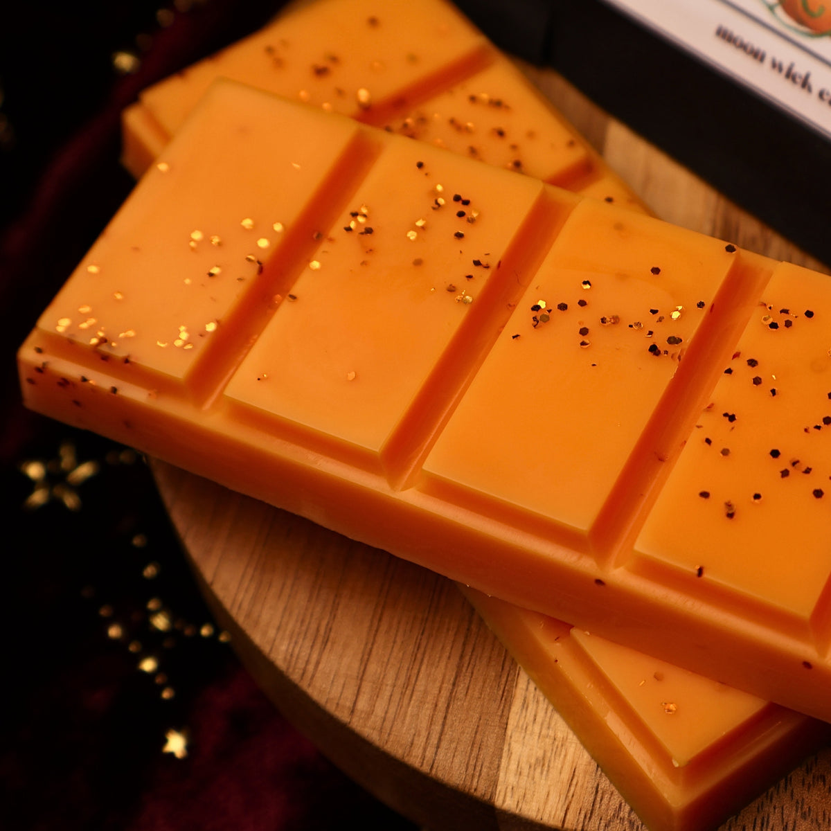 An orange wax melt snap bar with a segmented rectangular design, topped with orange glitter resting on a wooden riser.