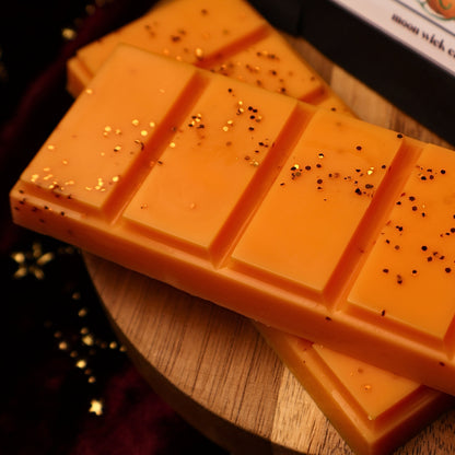 An orange wax melt snap bar with a segmented rectangular design, topped with orange glitter resting on a wooden riser.
