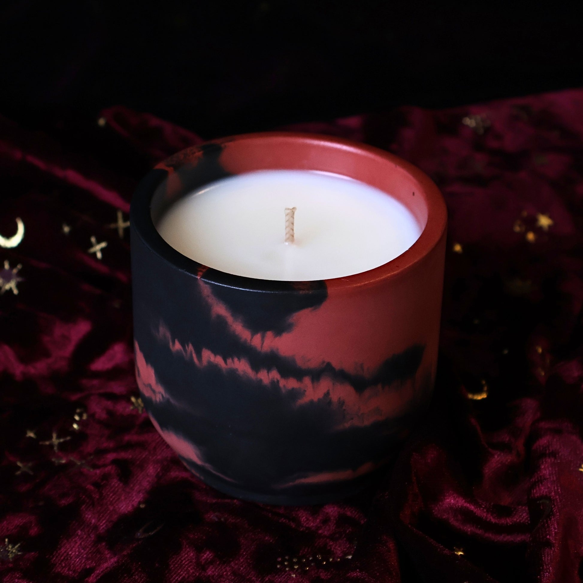 A marbled black and dark red hydrostone candle jar with a white wax fill, set against a dark background and draped deep burgundy velvet fabric with subtle gold star details.