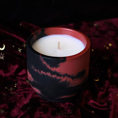 A marbled black and dark red hydrostone candle jar with a white wax fill, set against a dark background and draped deep burgundy velvet fabric with subtle gold star details.