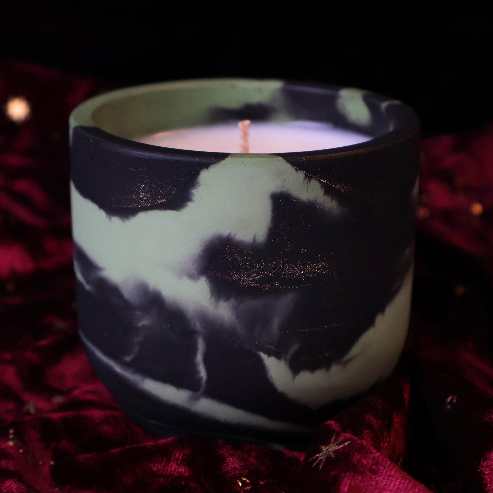 A light green and black marbled hydrostone container candle with gold glitter accents against a burgundy velvet fabric with celestial details